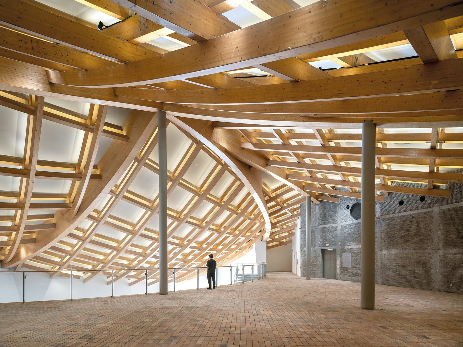 Interno di un edificio contemporaneo caratterizzato da una complessa struttura portante in legno, composta da grandi travi curve e intrecciate. Il pavimento è in mattoni e alte colonne sostengono la copertura; una persona osserva lo spazio da una passerella interna.