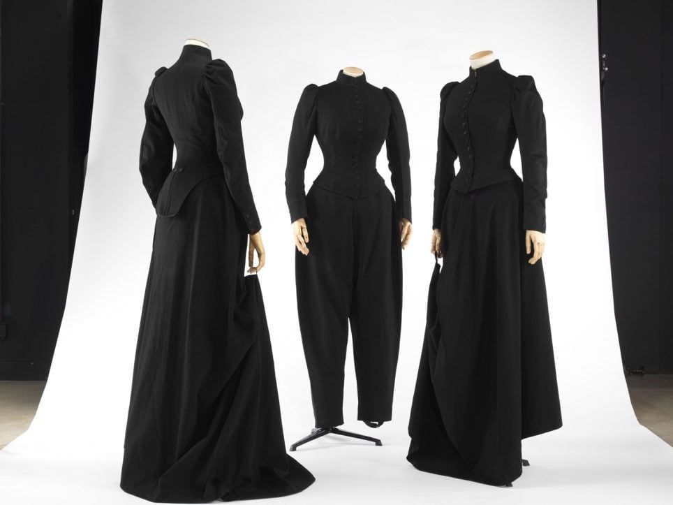 Tre abiti femminili neri di foggia storica esposti su manichini, caratterizzati da corpetti aderenti, maniche lunghe e spalle strutturate. Due presentano lunghe gonne, mentre quello al centro è abbinato a pantaloni ampi.