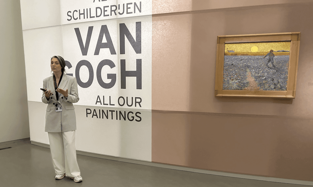 Interno di una sala espositiva dedicata a Vincent van Gogh. Una donna parla davanti alla scritta “Van Gogh – All Our Paintings”; sulla parete accanto è esposto un dipinto raffigurante un seminatore in un campo al tramonto.