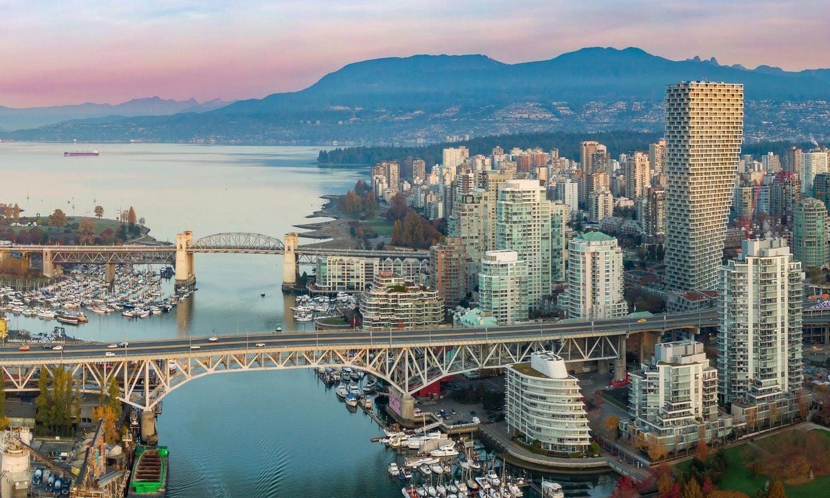 Veduta panoramica di Vancouver, con numerosi grattacieli affacciati sull’acqua, porti turistici e due ponti in primo piano. Sullo sfondo si estendono la baia e le montagne, sotto un cielo dalle tonalità rosa e azzurre.