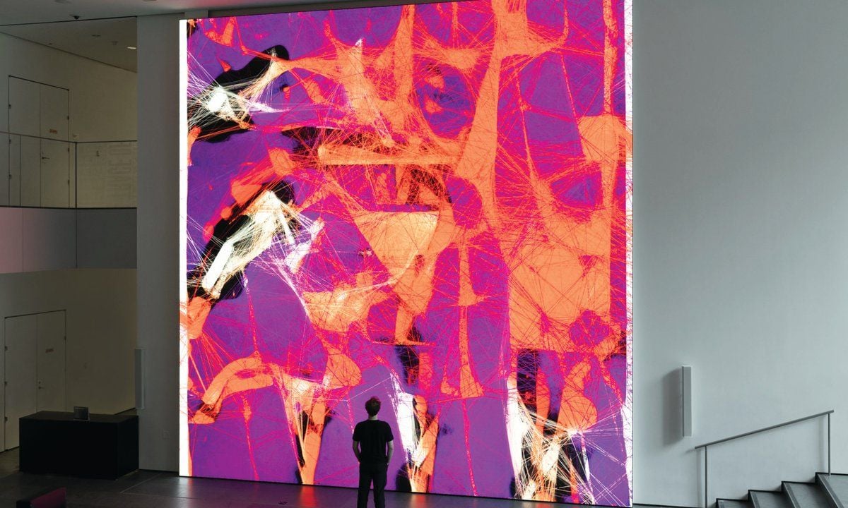 Interno di uno spazio espositivo con una grande installazione digitale dai colori viola, rosa e arancione, composta da figure e intrecci di linee sovrapposte. Una persona osserva l’opera dal basso, evidenziandone le grandi dimensioni.