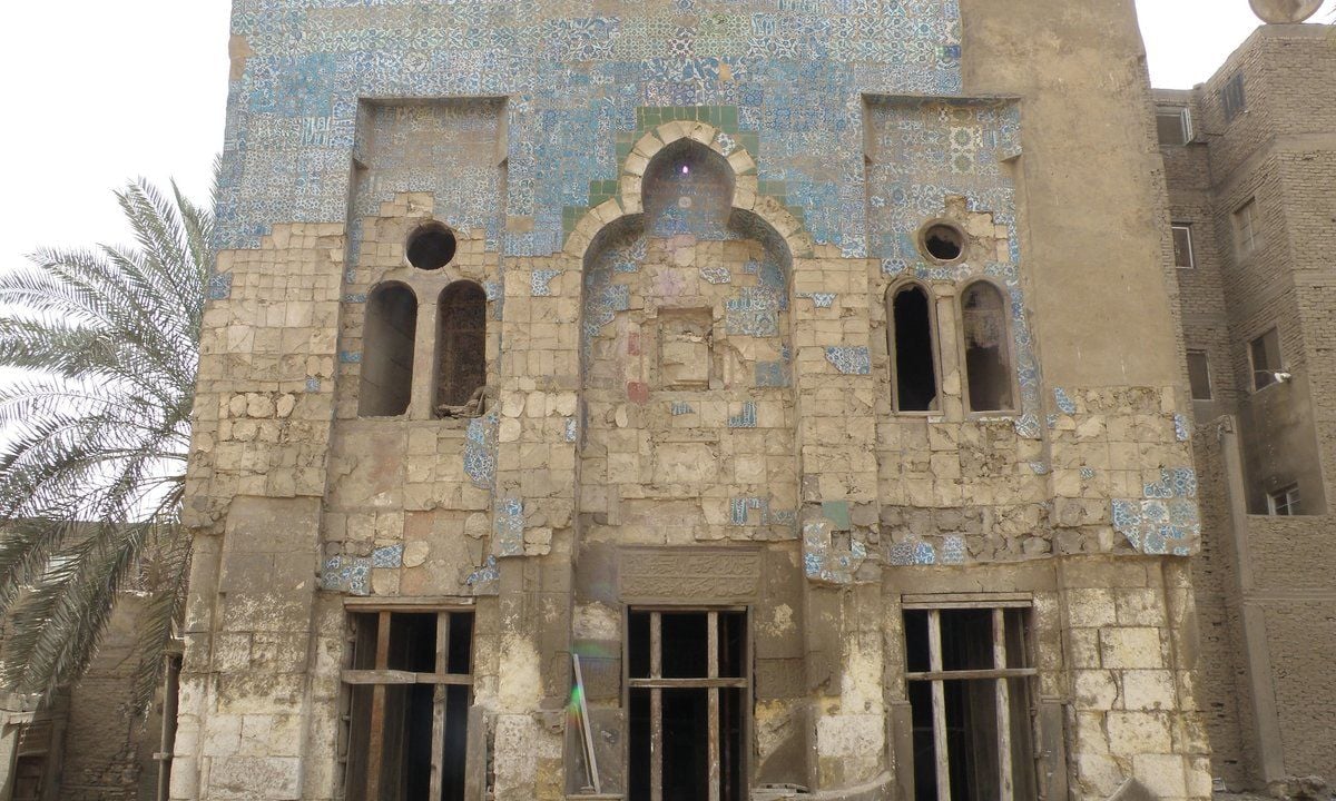 Facciata di un edificio storico in pietra, parzialmente rivestita da piastrelle decorative azzurre. La struttura presenta archi e finestre di forme diverse e mostra segni di deterioramento e parti mancanti.