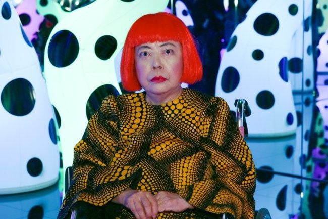 The artist Yayoi Kusama sitting in a wheelchair inside a room dedicated to her works.